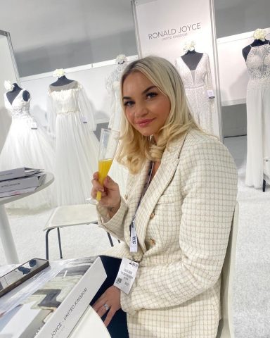 A woman with long blonde hair holding a yellow drink, seated near wedding dresses.